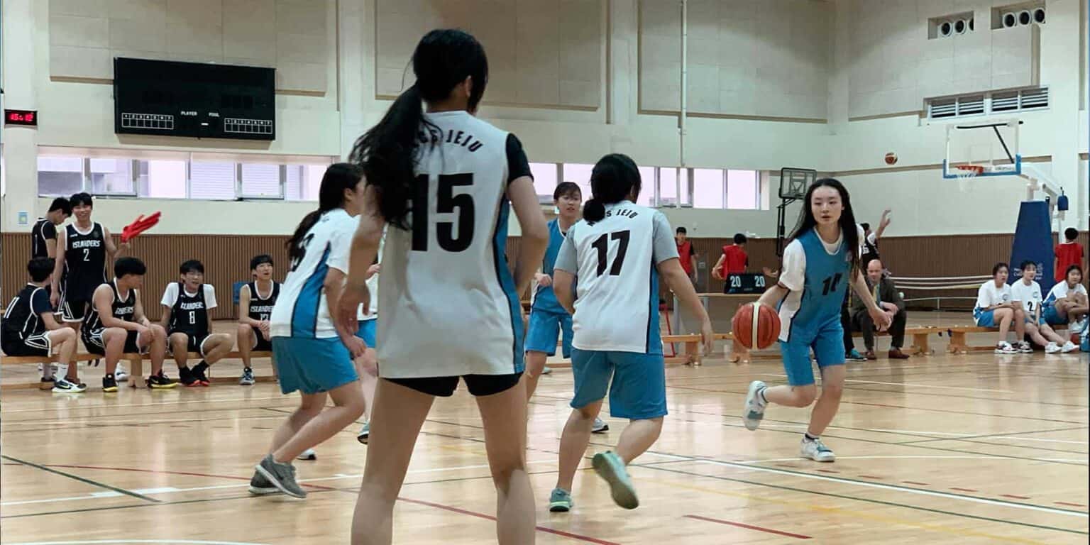 Young female basketball players during a game at an indoor school gymnasium with spectators.