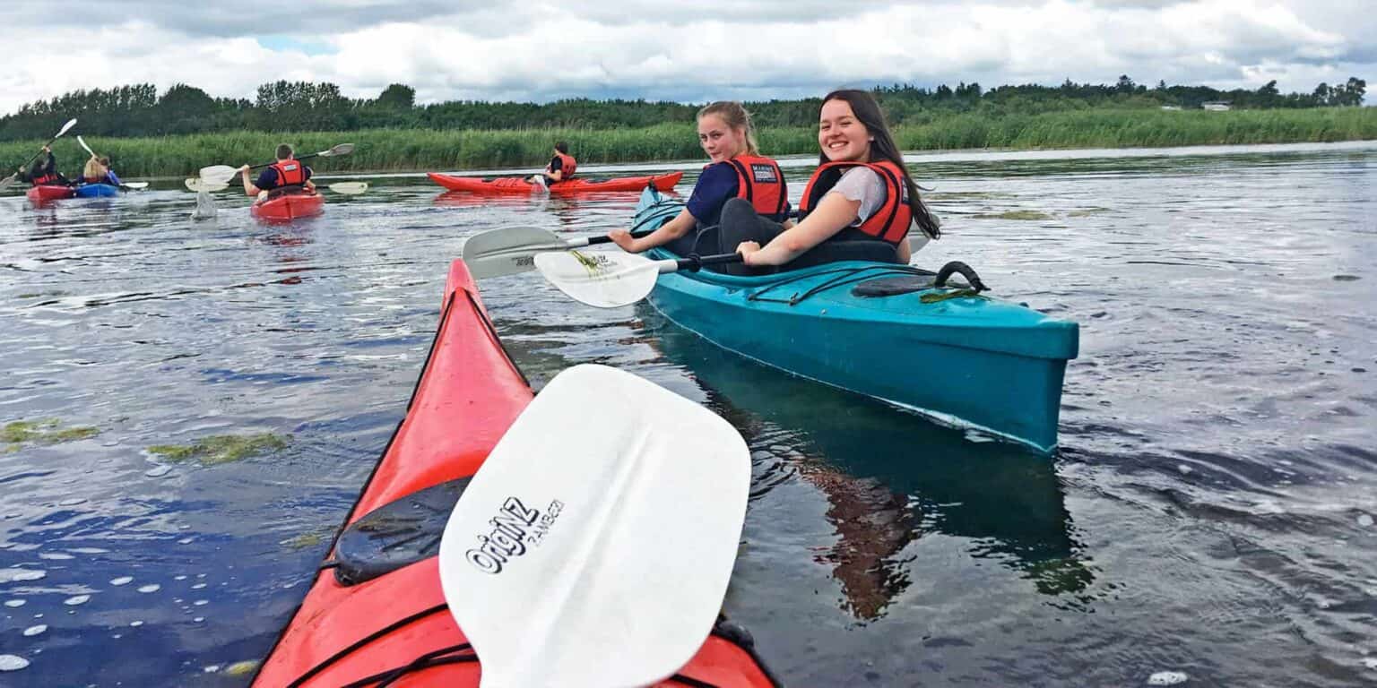 Children kayaking on a calm river with lush greenery in the background, outdoor water activity at a school camp.