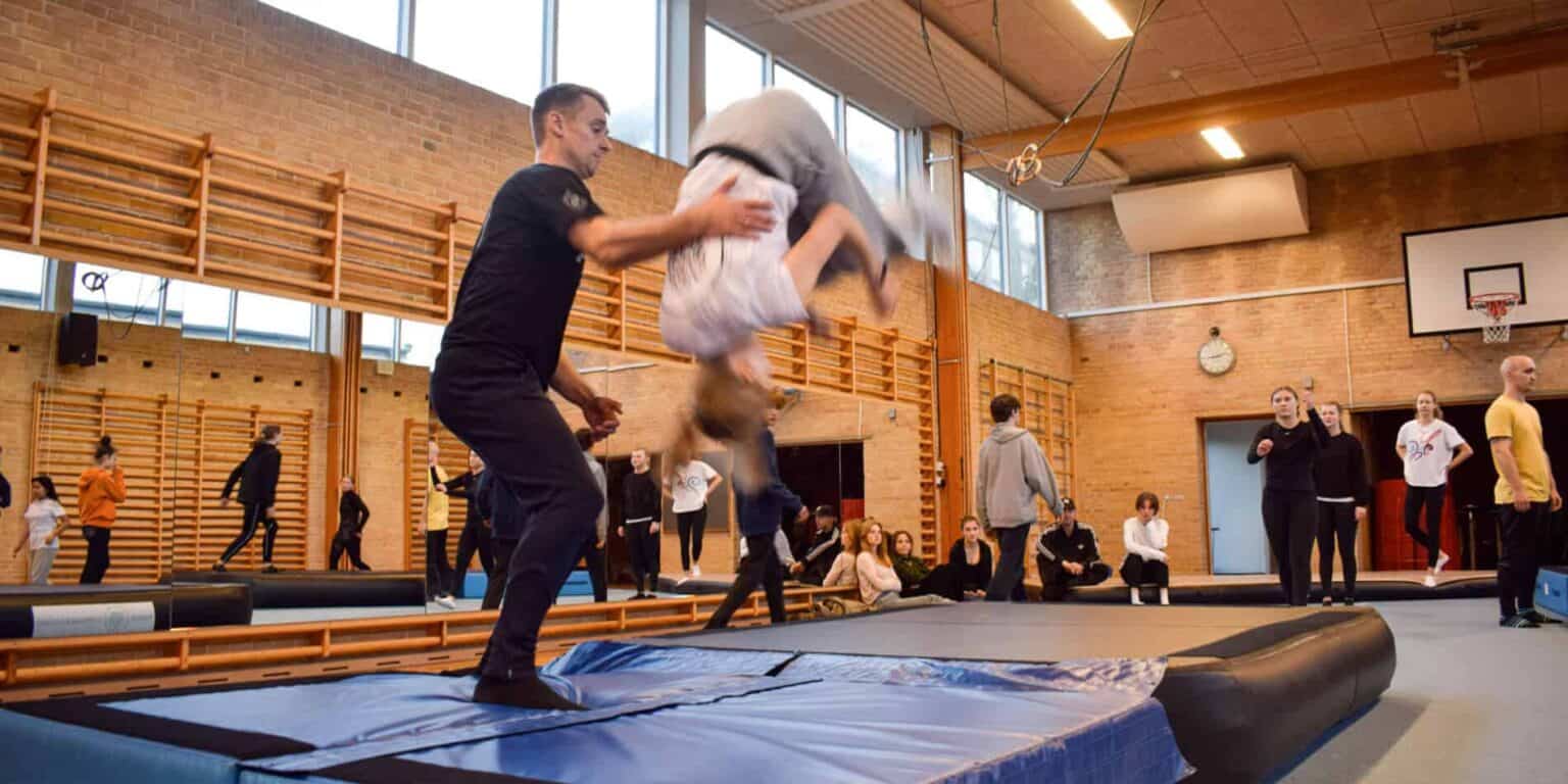 Children practicing gymnastics on a trampoline in a school gymnasium with coaches and peers.