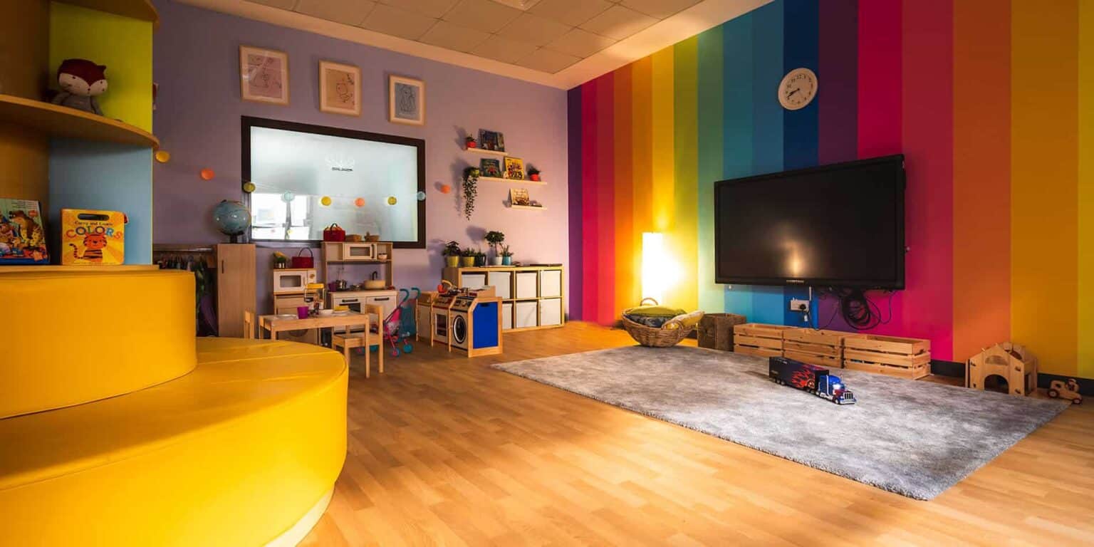 Aula moderna y colorida con decoración de arco iris en las paredes, juguetes educativos y acogedoras zonas de lectura y juego para los niños.