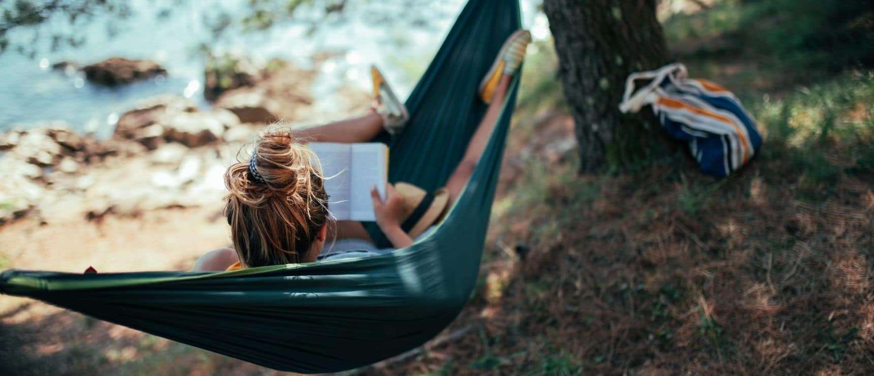 Relaxed student reading outdoors in hammock by lake, enjoying nature for stress relief and improved focus.