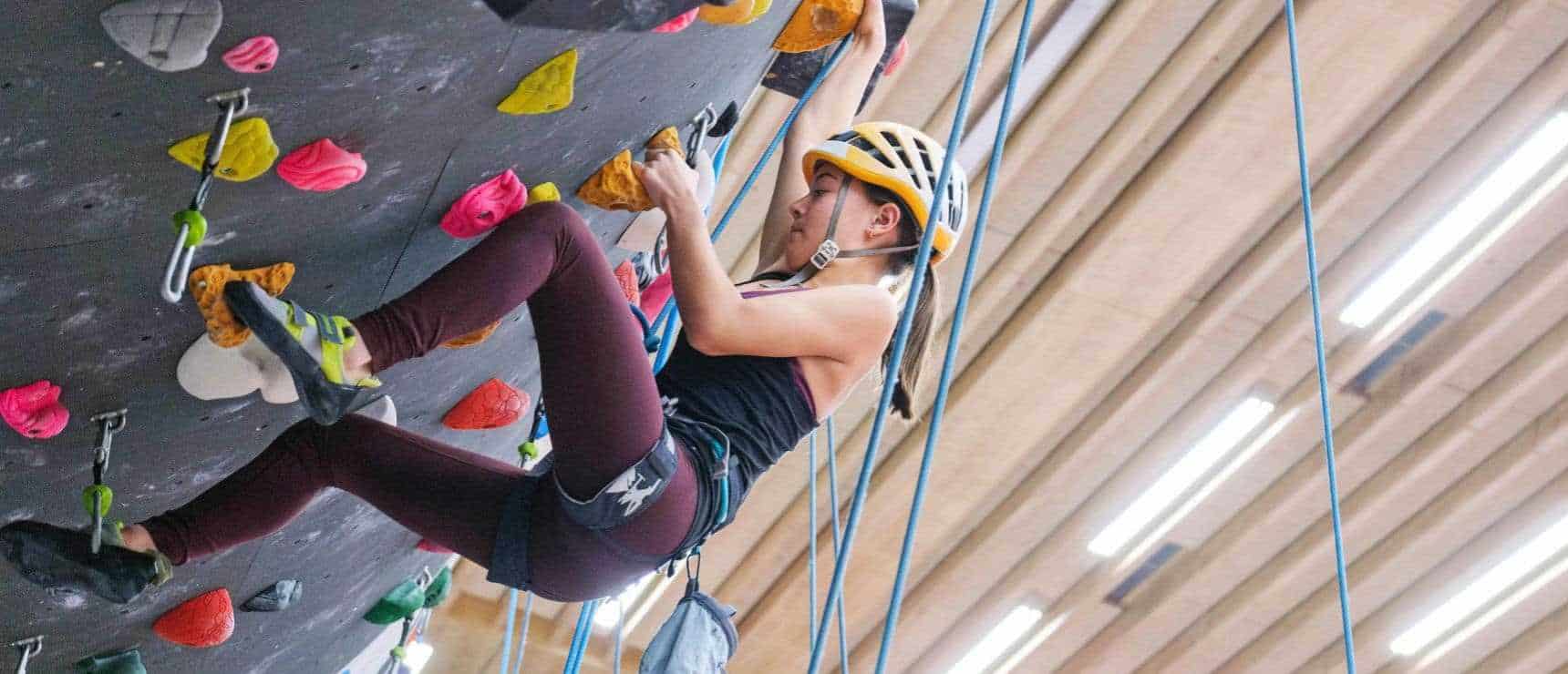 Young girl rock climbing indoors at a school gym, wearing safety helmet and harness.