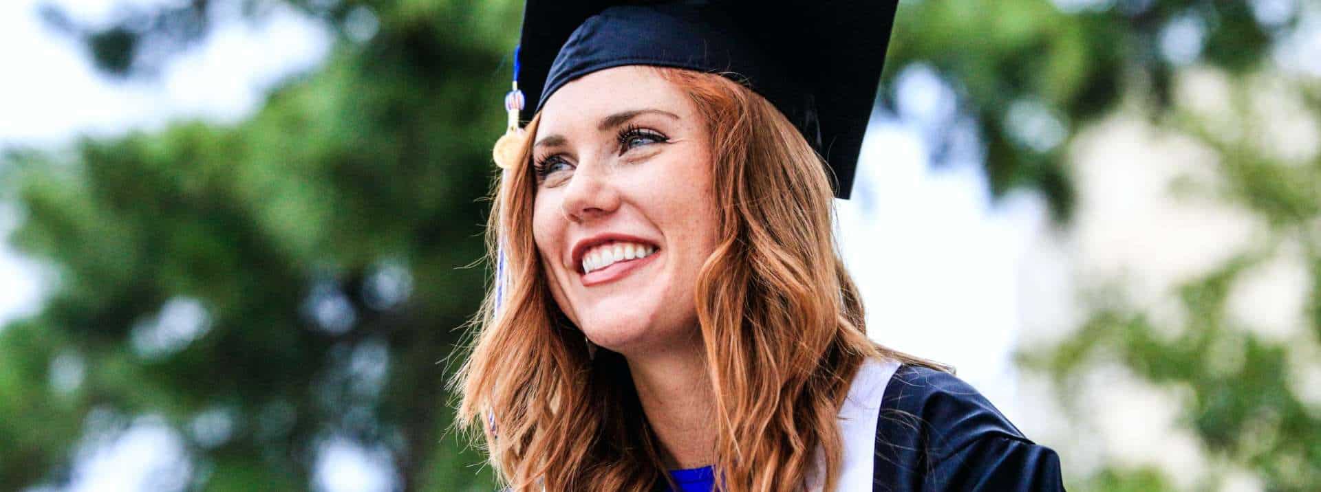 Bright smiling female graduate in cap and gown celebrating outdoor graduation ceremony.