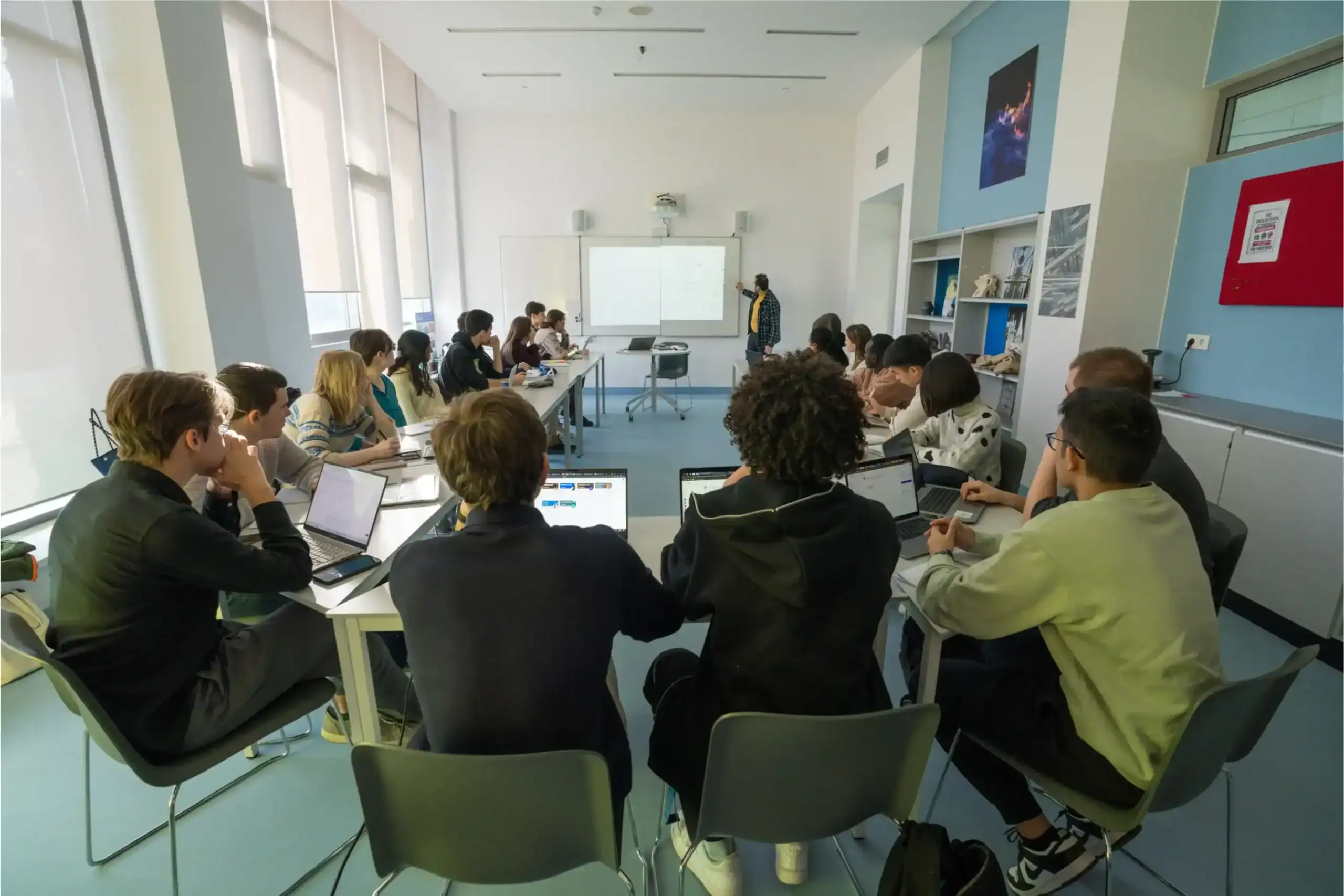 Students in a modern classroom at World Schools with laptops and a teacher presenting at the whiteboard.