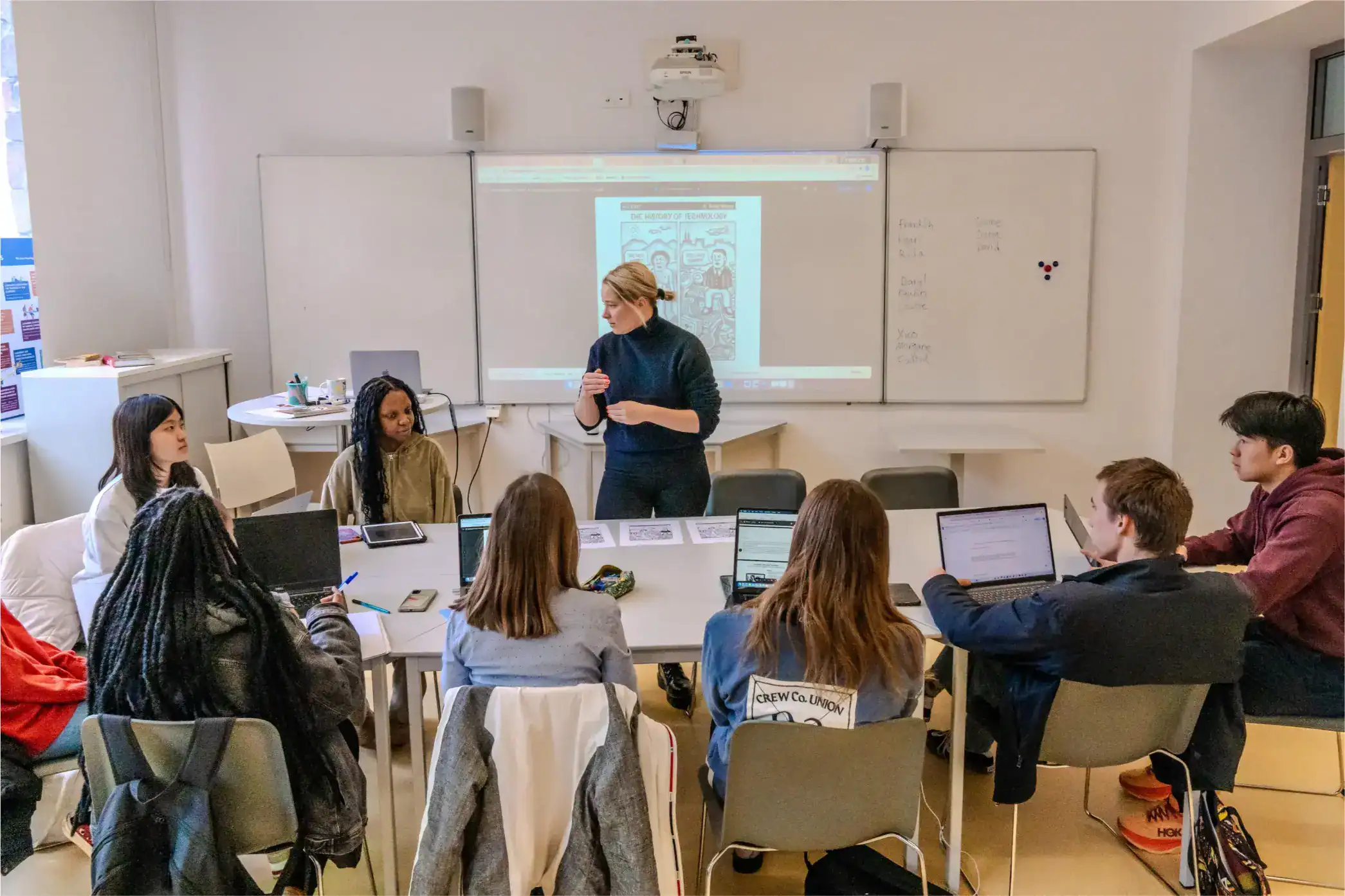 Students in a modern classroom attending a lesson with a teacher presenting on a digital smartboard.