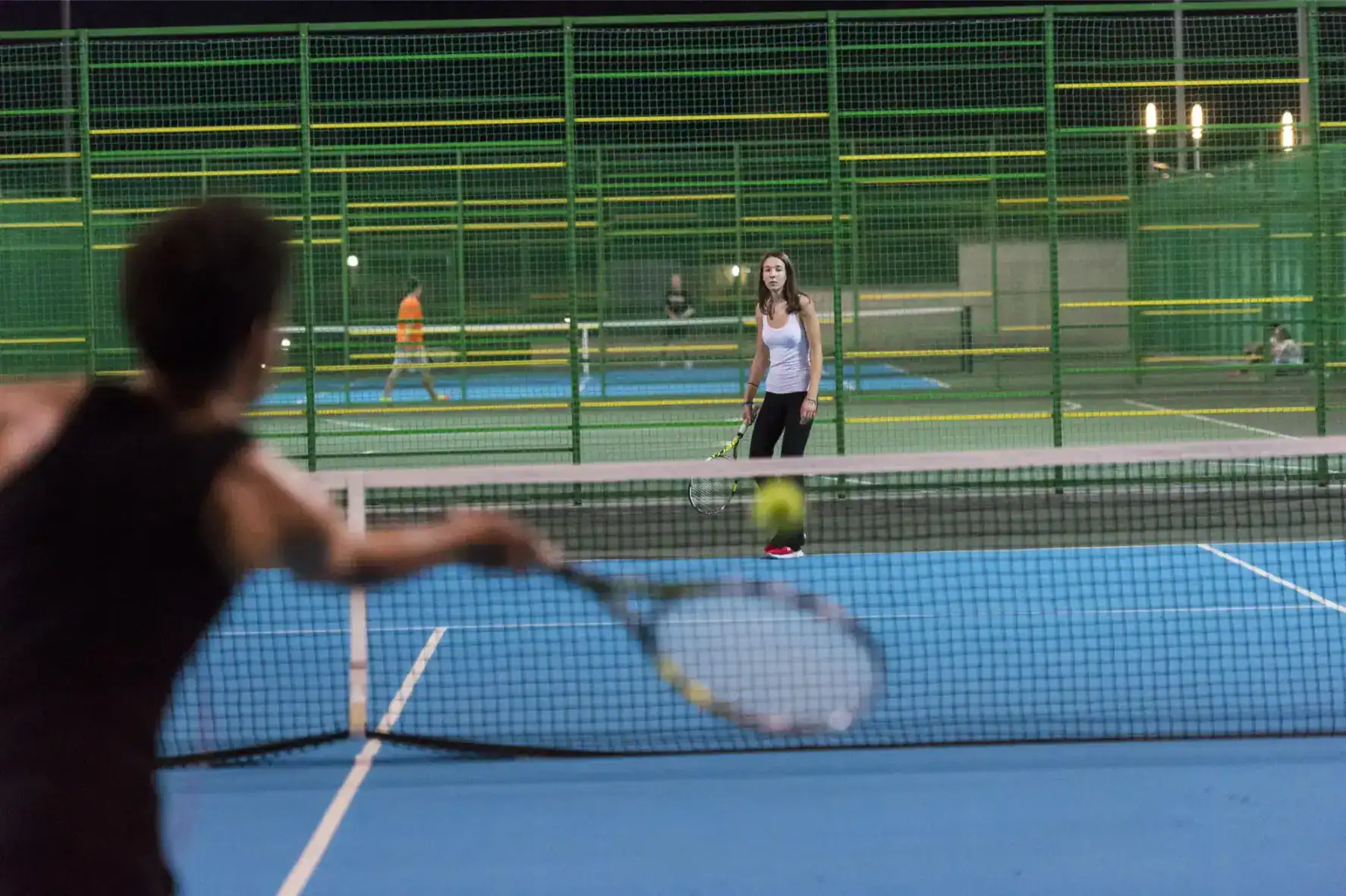 Tennis match at night on an outdoor court with green fencing, featuring young players and a focus on sports education at international schools.