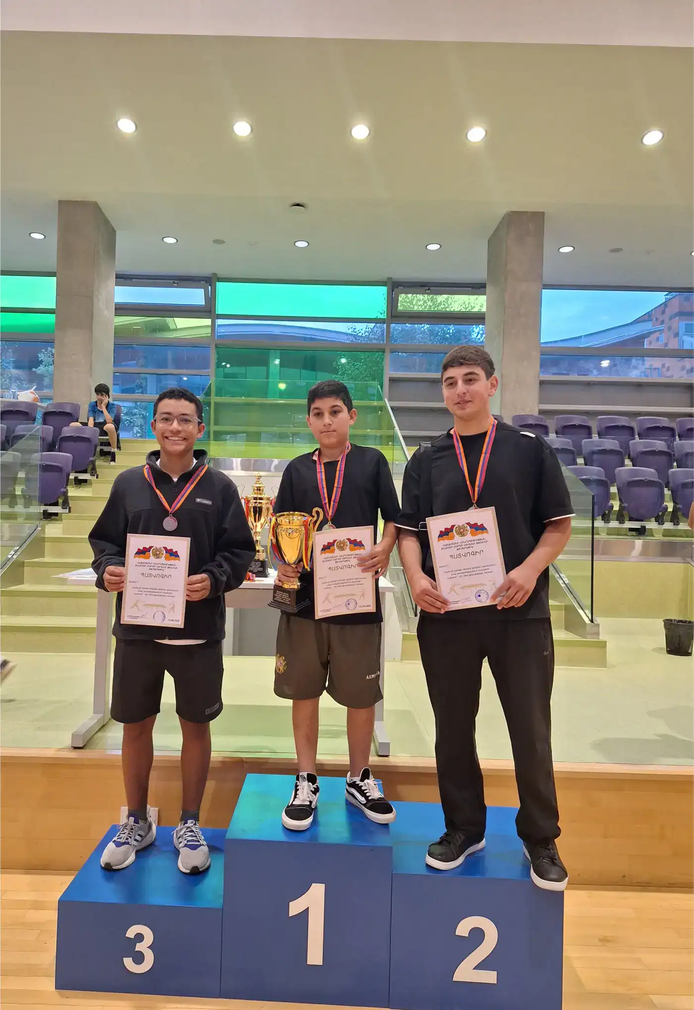 Three young students standing on winners' podium with medals, certificates, and trophies at a school competition.