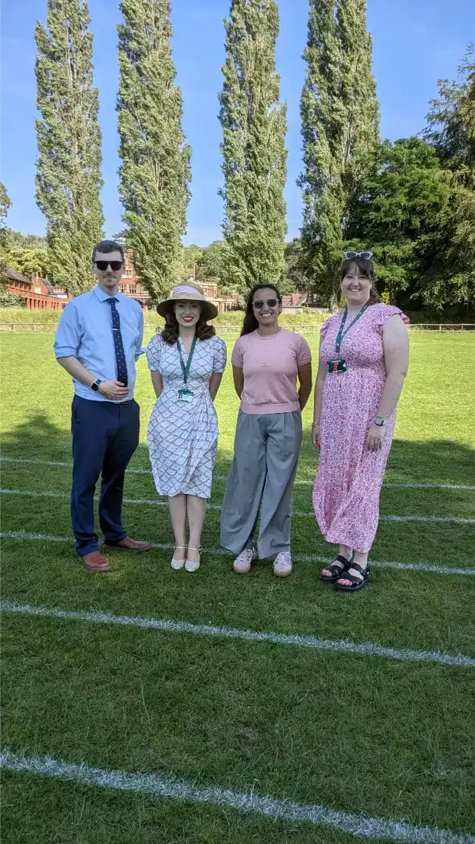 Group of four students standing on a sports field with tall trees in the background.