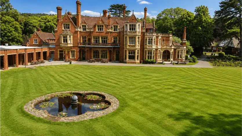 Historic mansion with lush green lawn and pond in front, surrounded by trees.