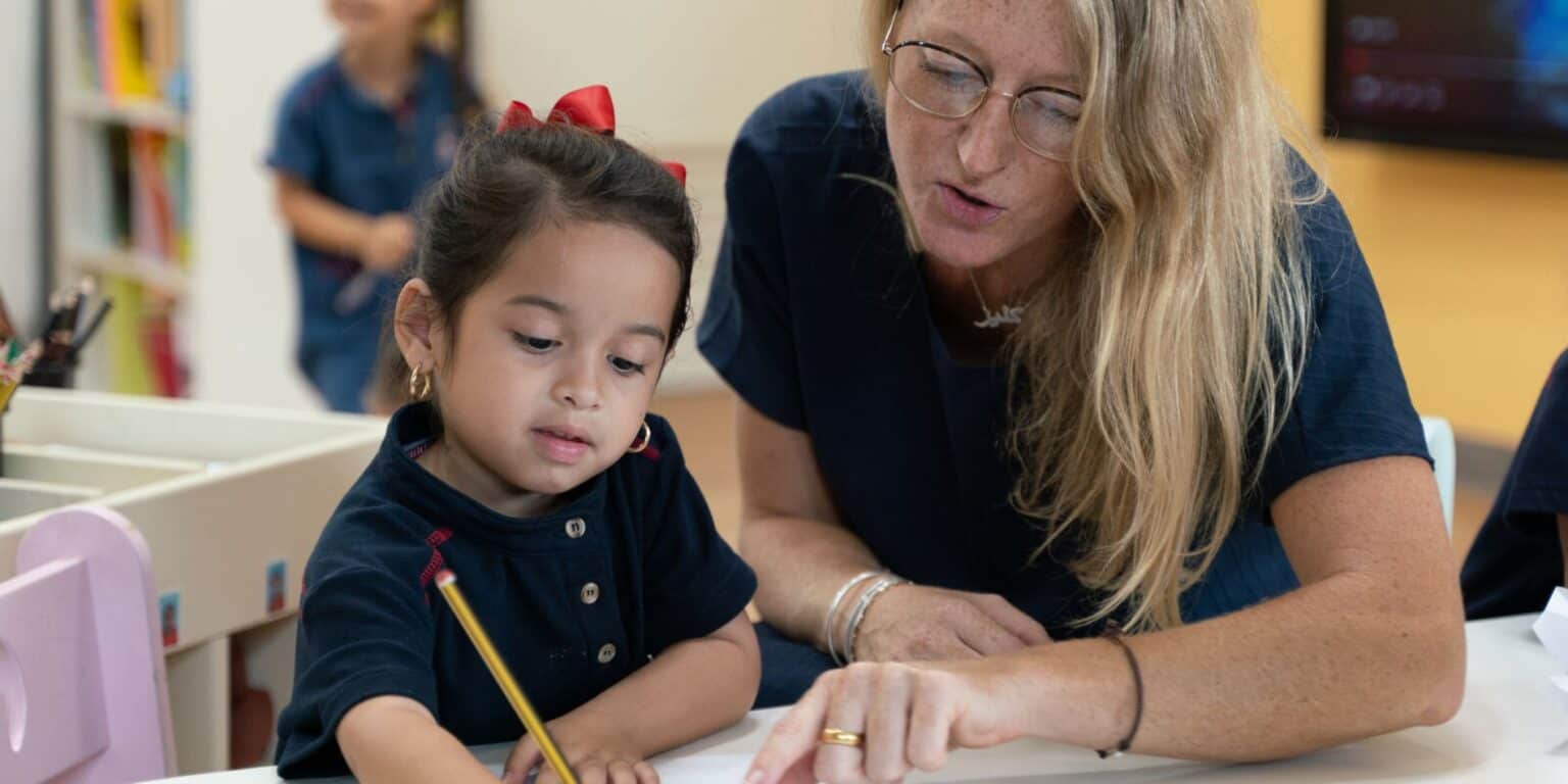 Focused teacher assisting young girl with schoolwork in a classroom setting.