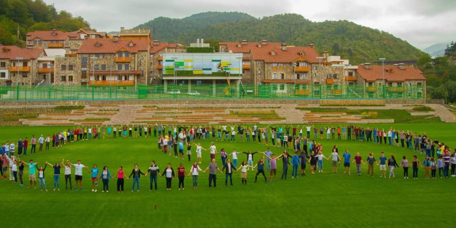 Children holding hands in a circle on a lush green field at a global school campus.