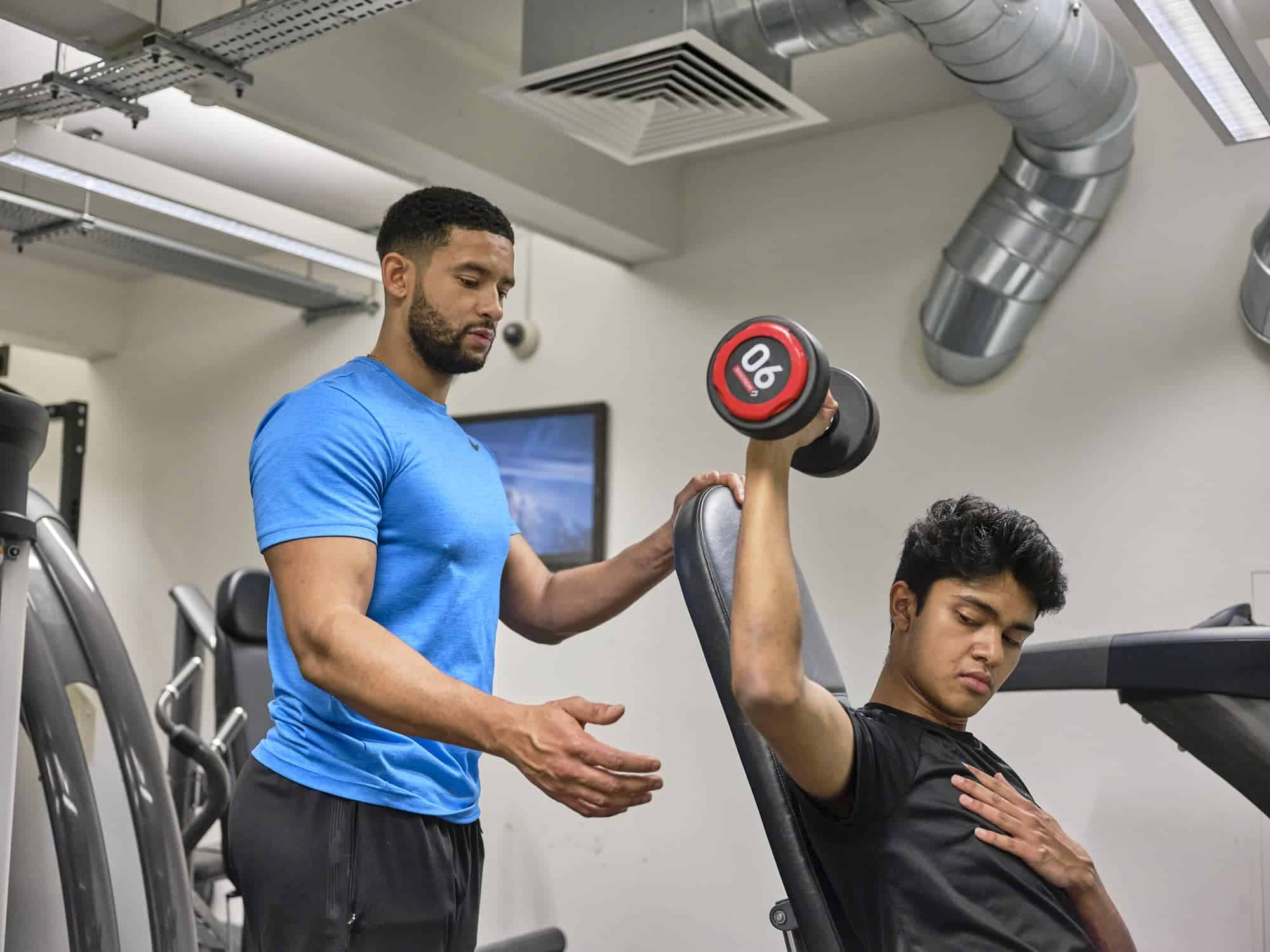 Personal trainer assisting young man with dumbbell exercise at gym.