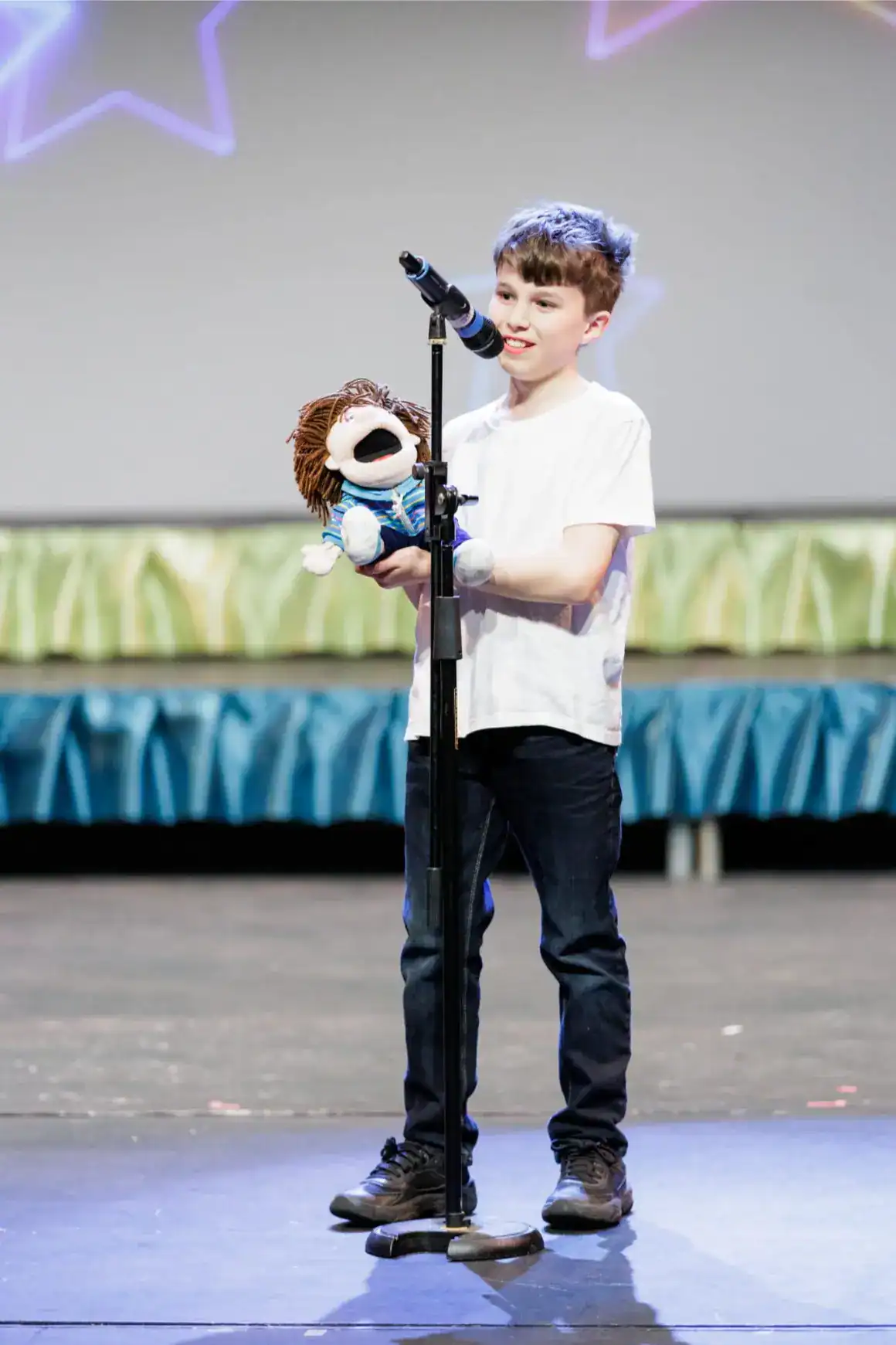 Young boy performing on stage with puppet in a school talent show, showcasing creativity and confidence.