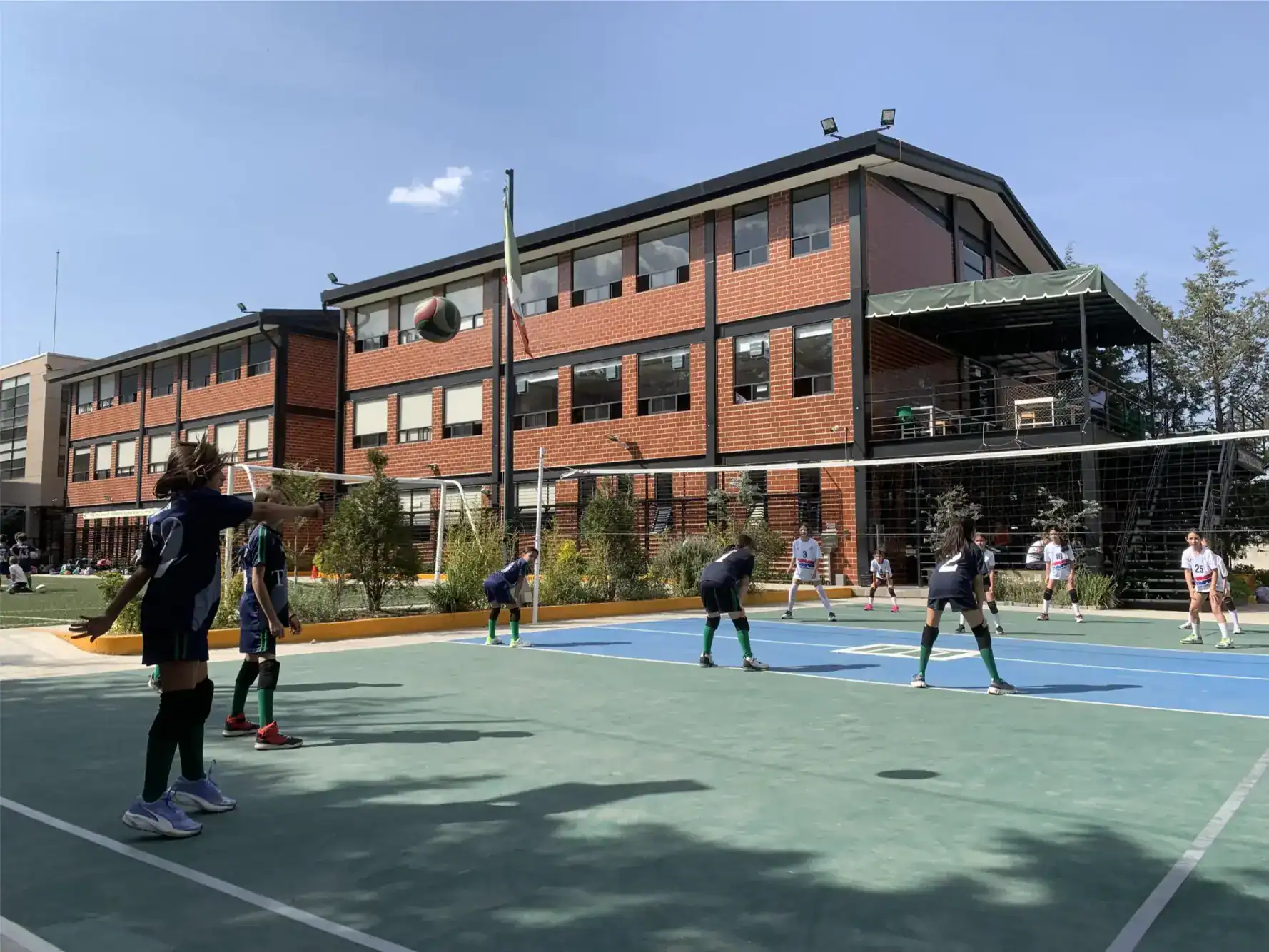 Students playing volleyball at a school outdoor sports court with a brick school building in the background.