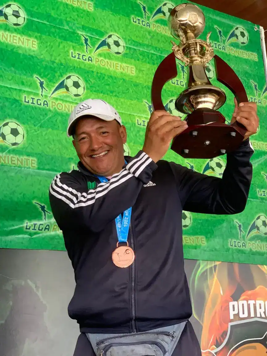 Victor trophy celebration at Liga Poniente football match, smiling coach holding up golden trophy on green backdrop.