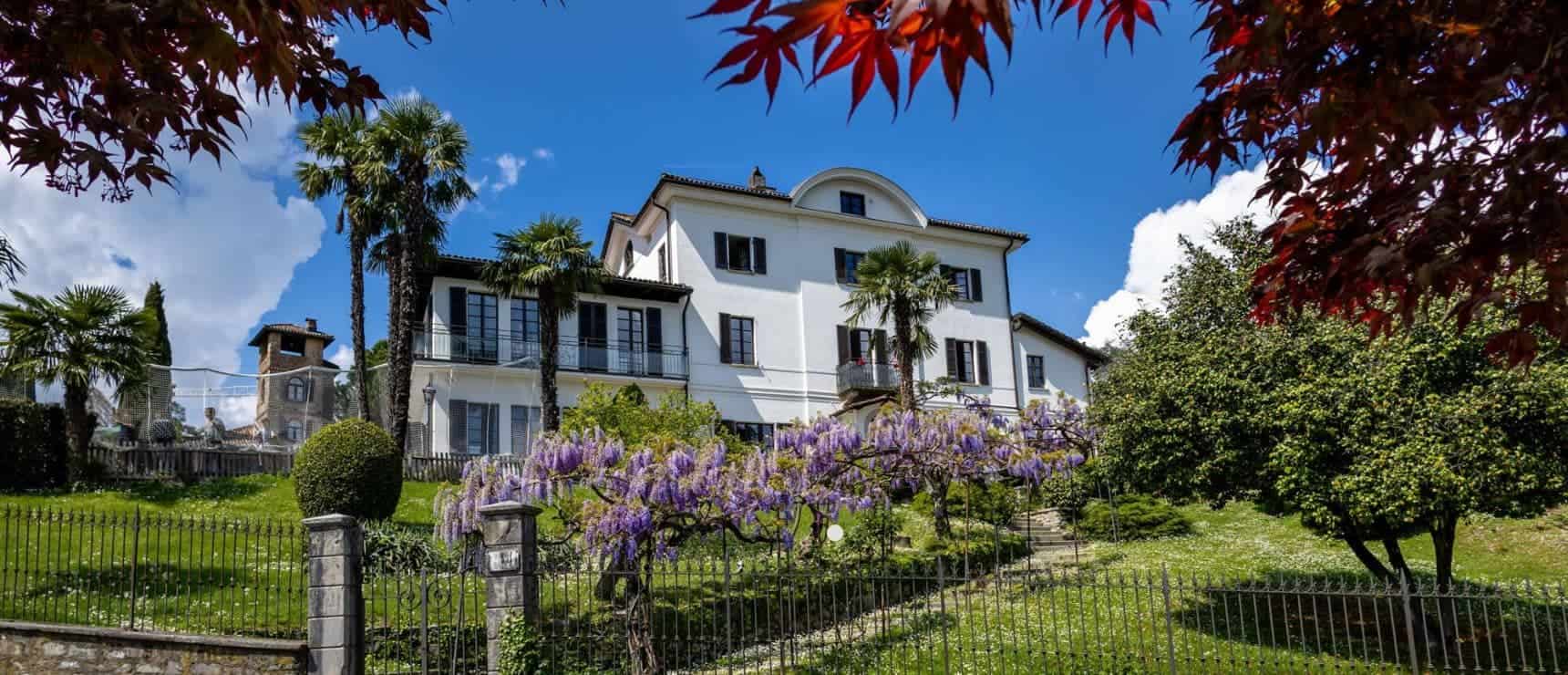 Luxurious white mansion surrounded by lush gardens and palm trees, in sunny weather.