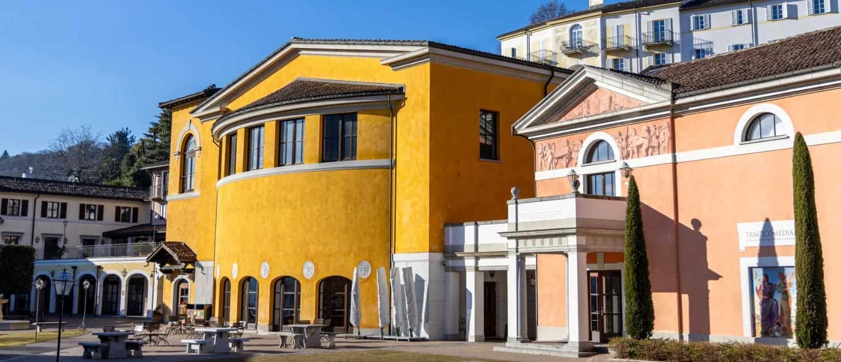 Colorful European-style school building with vibrant yellow and orange facades, surrounded by outdoor seating and tall cypress trees.