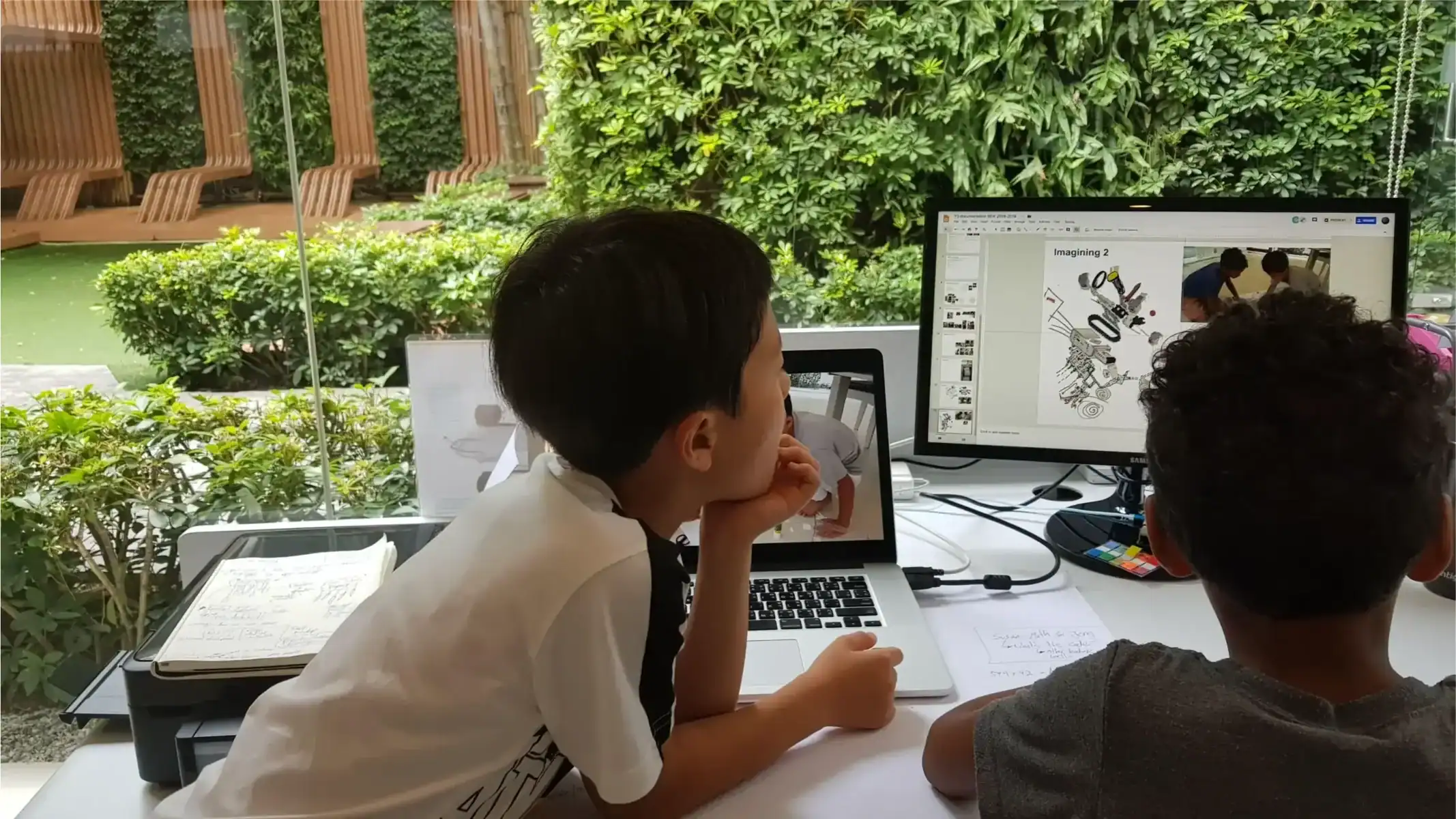 Students working on digital design projects in a modern classroom with greenery outside.
