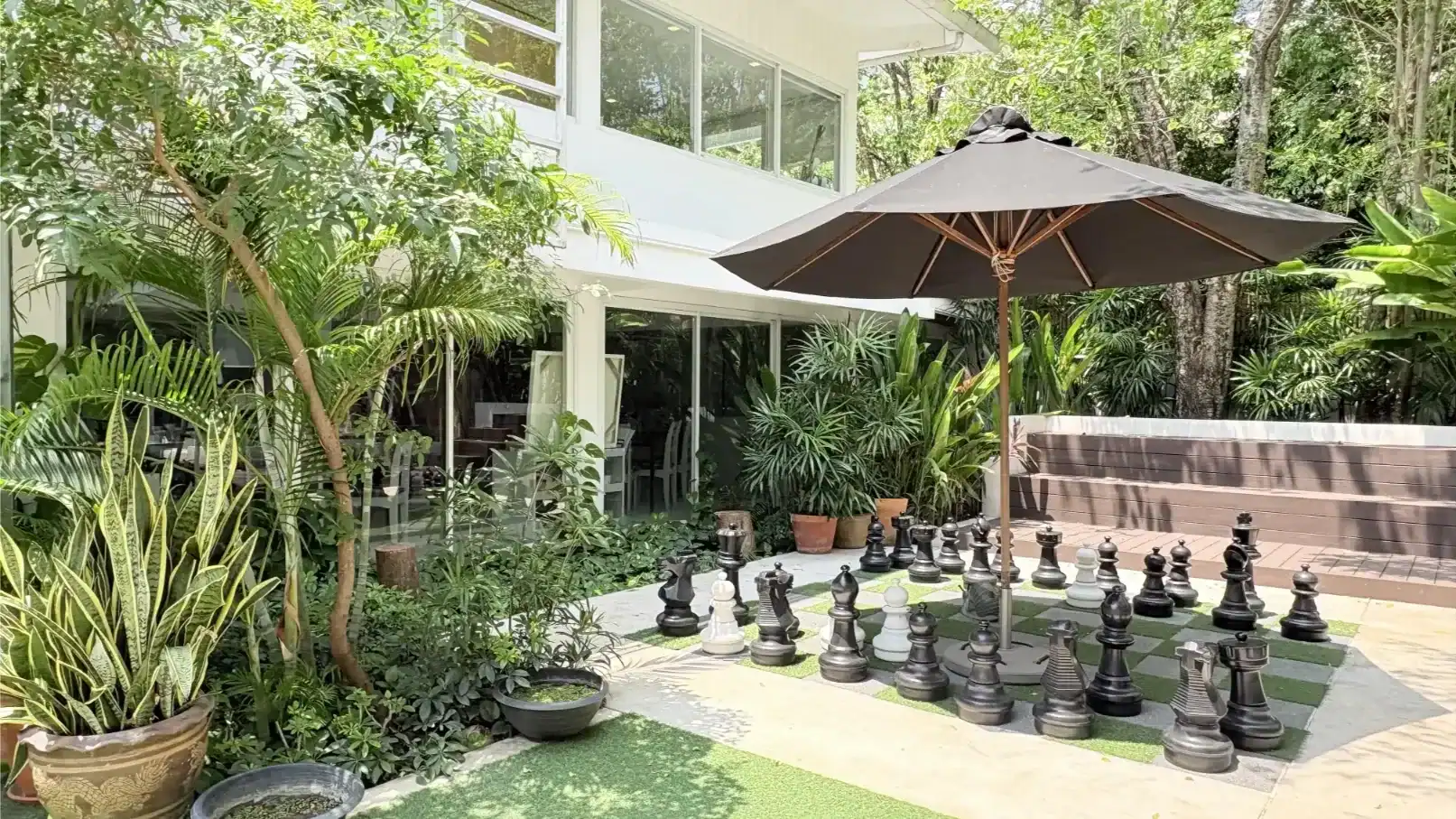 A lush outdoor garden with a large chess set and seating area under a black umbrella.