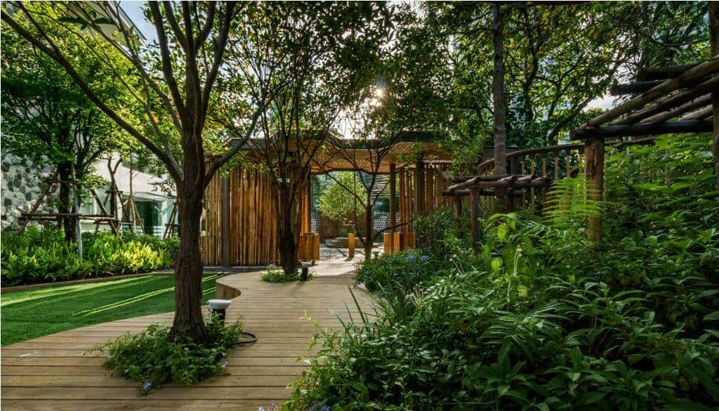 A lush green garden with wooden pathways and a gazebo, surrounded by trees and tropical plants.