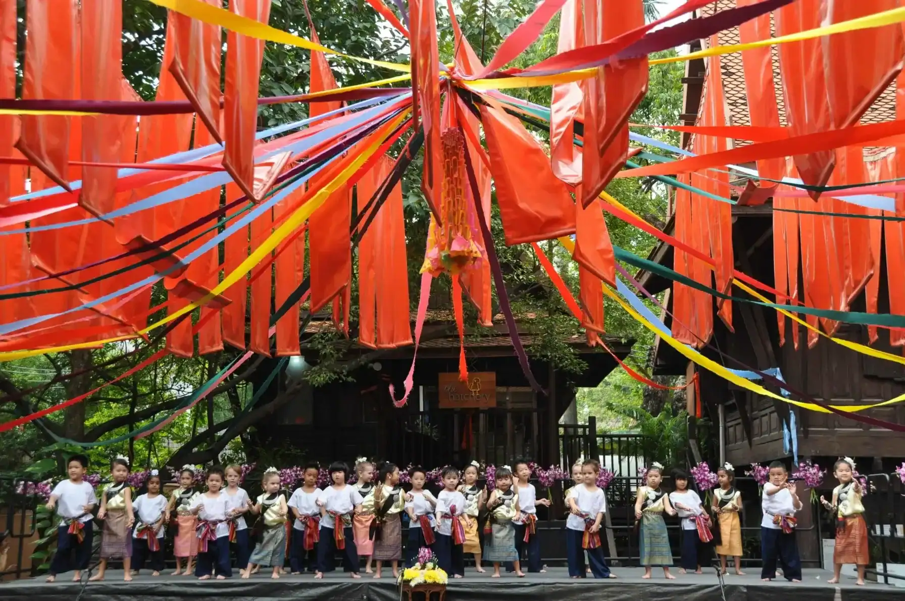 A colorful traditional festival with children performing on stage under vibrant hanging decorations.