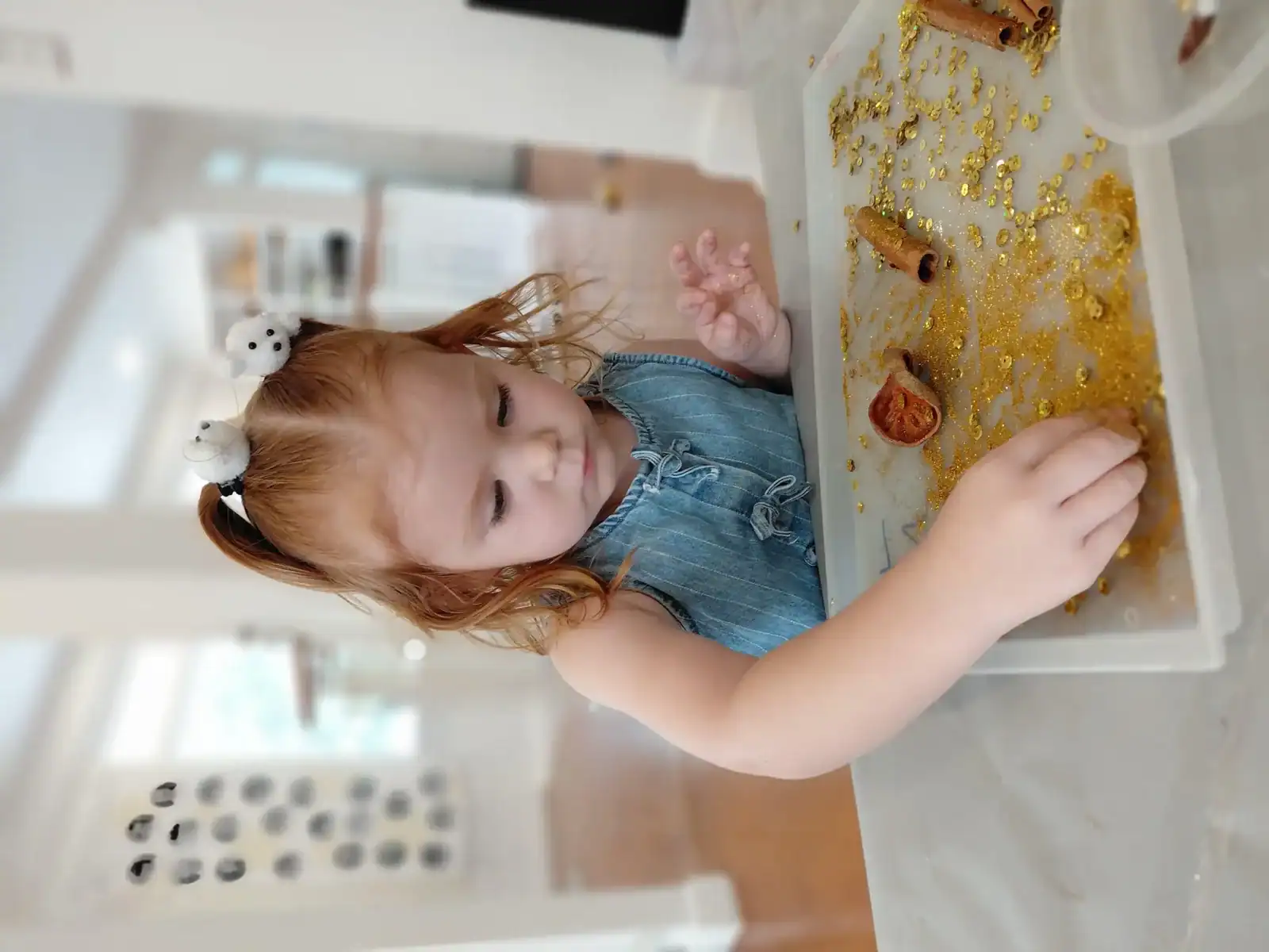 Young girl enjoying sensory play with golden glitter and cinnamon sticks at home.