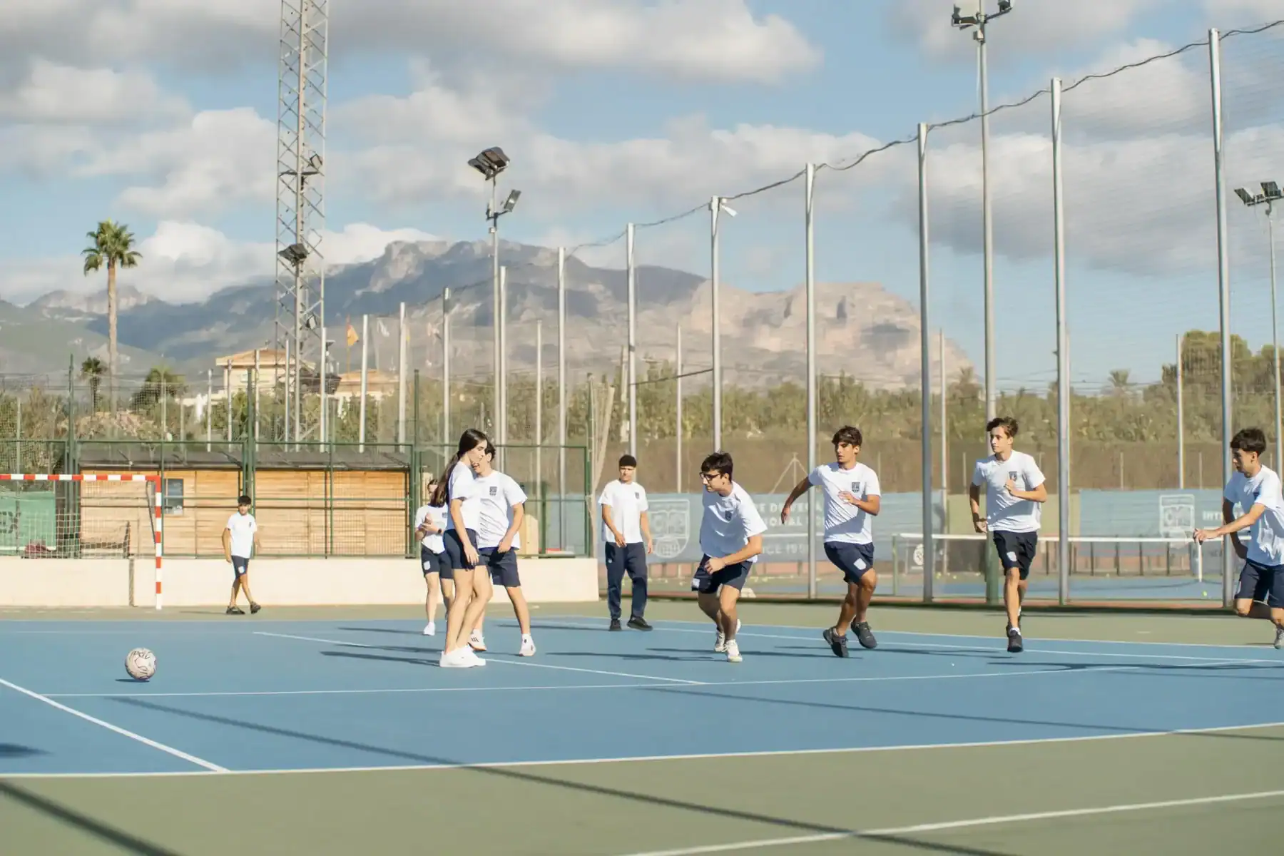 Students playing soccer on outdoor sports court at World Schools campus with scenic mountains in background.