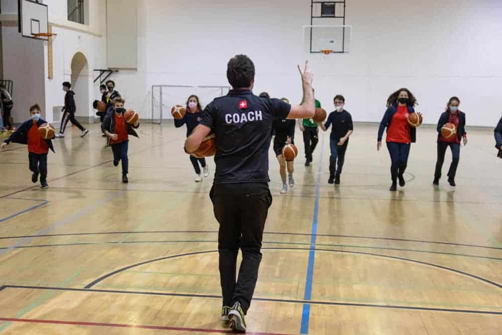 Motivational coach instructing children in basketball practice at a school gymnasium.