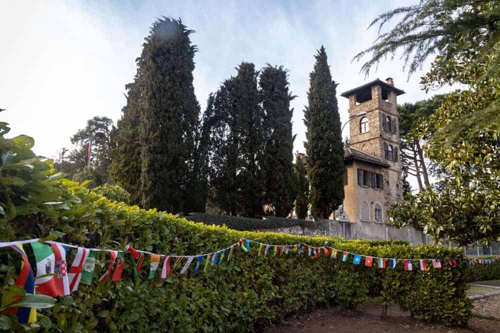 Colorful international flags decorate a lush green hedge in front of a historic stone building with tall cypress trees.