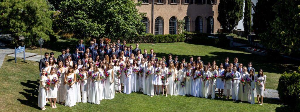Graduating students in white dresses and suits holding flowers outside a historic school building.