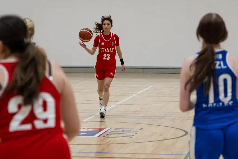 This Month at TASIS: January 2025 10 Basketball player in red jersey dribbling on indoor court with opponents in blue jerseys.