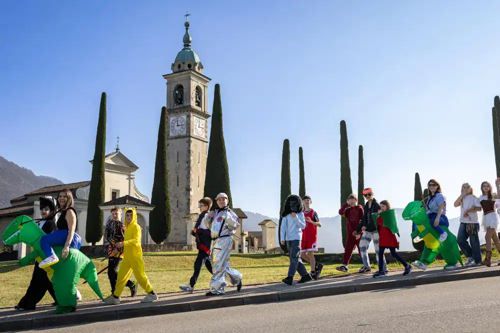 This Month at TASIS: March 2025 3 Colorful children parade dressed in costumes near a historic church with tall cypress trees, celebrating school events.