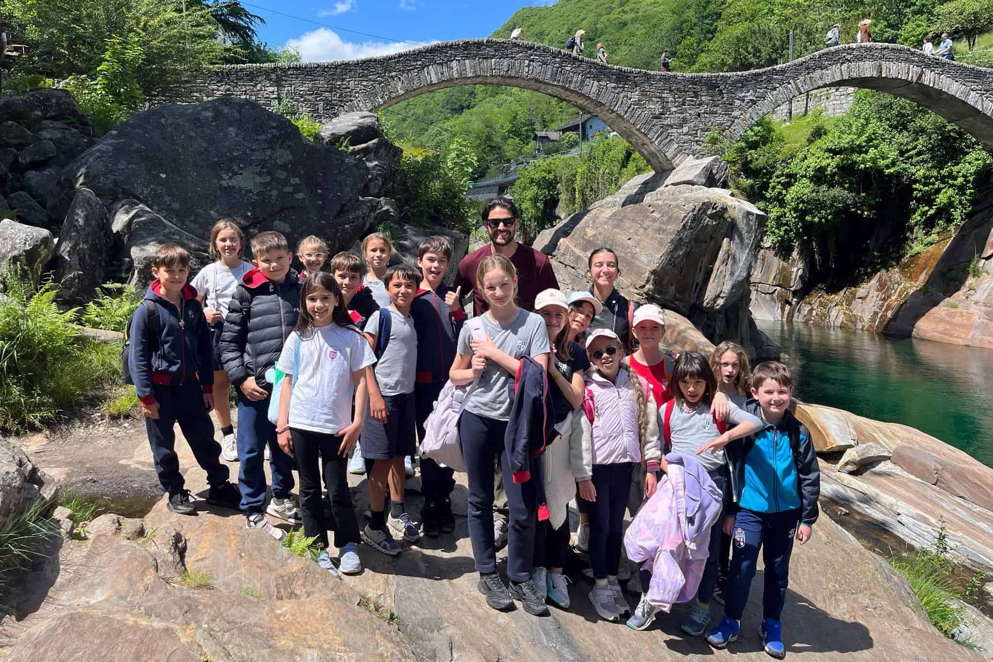 This Month at TASIS: May 2025 22 1. Group of diverse schoolchildren and teachers on nature trip at a scenic river with stone bridge.