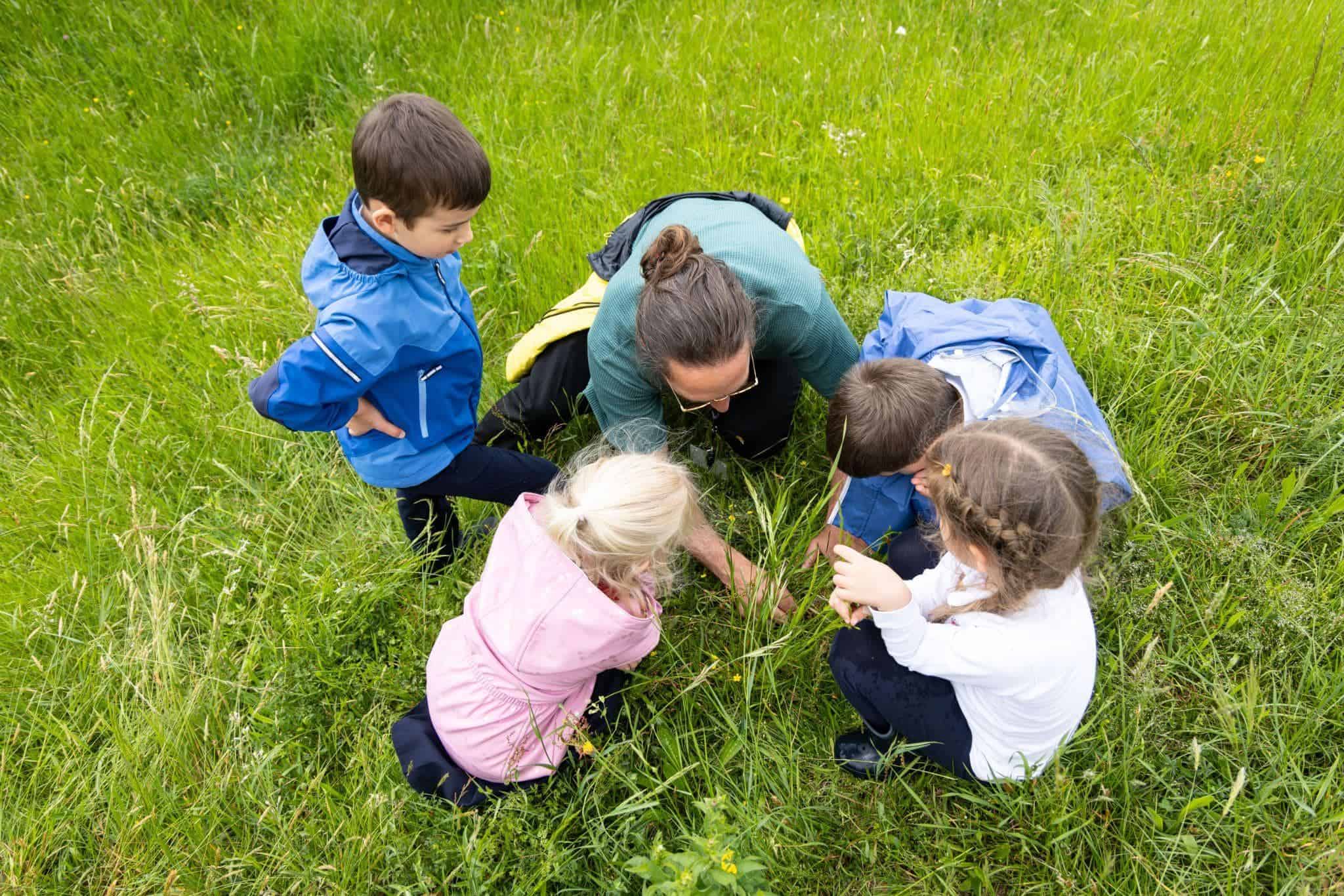 This Month at TASIS: May 2025 5 Children exploring nature with teacher outdoors in green field.