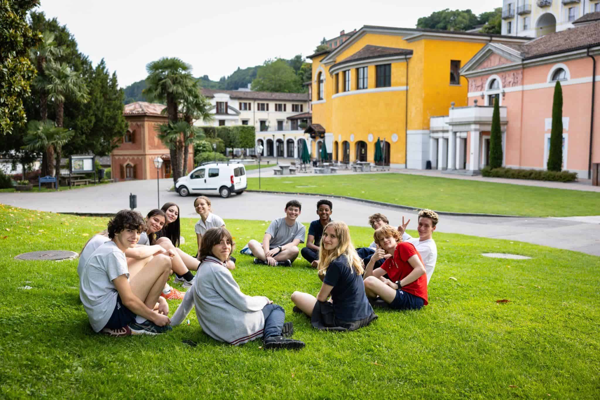 This Month at TASIS: May 2025 16 Bright students sitting on green grass outside a colorful international school campus.