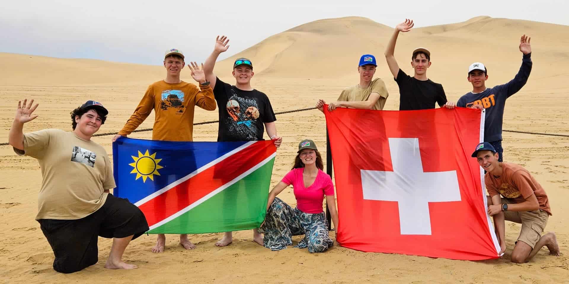 Bright students holding Namibia and Switzerland flags in desert landscape for world school exchange program.