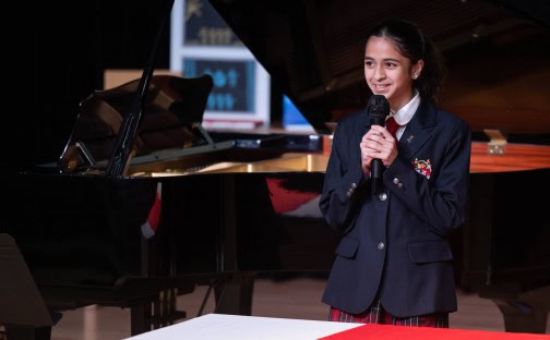 Young girl speaking at school event with microphone and grand piano in background.