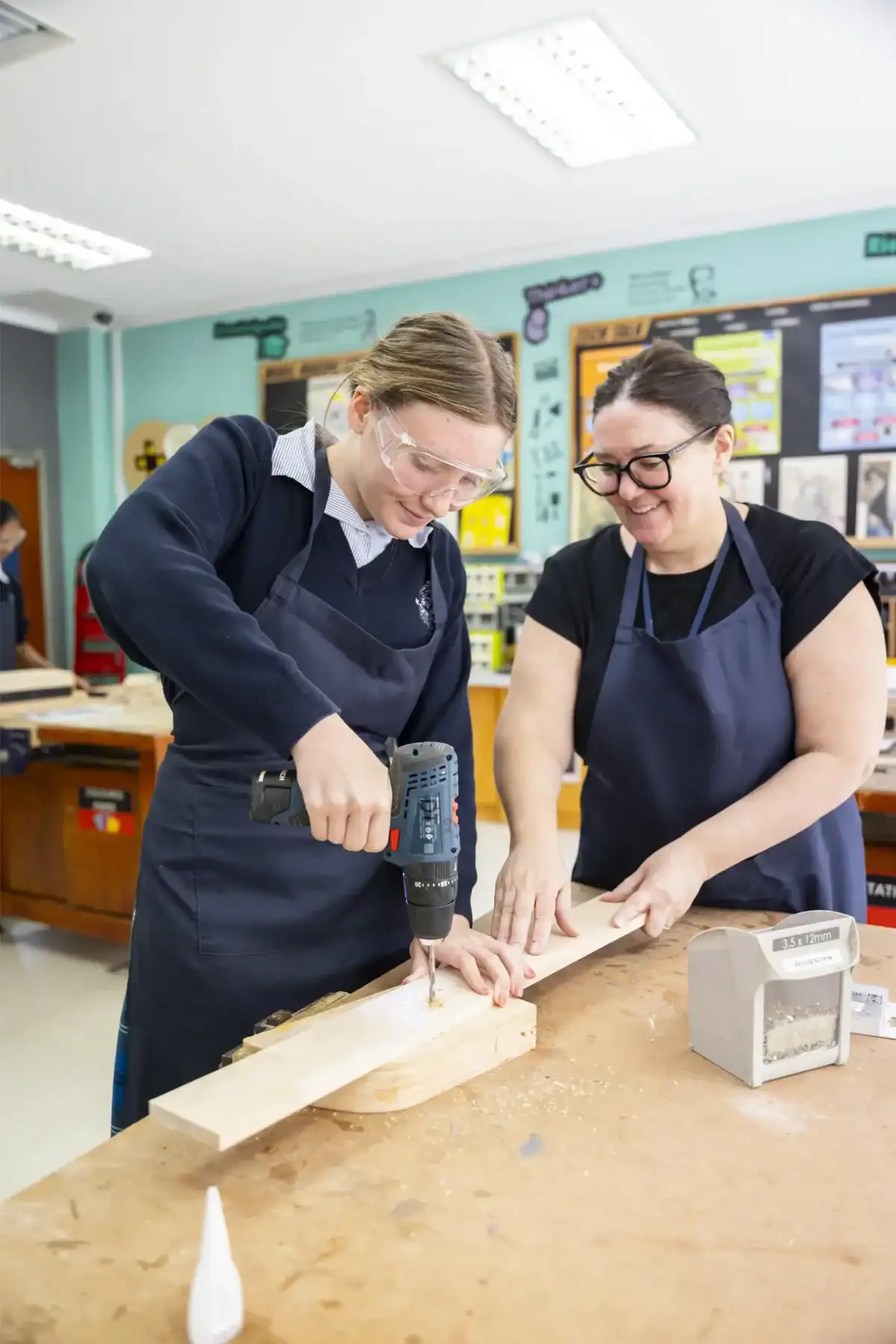 Two women working together on woodworking project in classroom, using power drill and safety glasses.