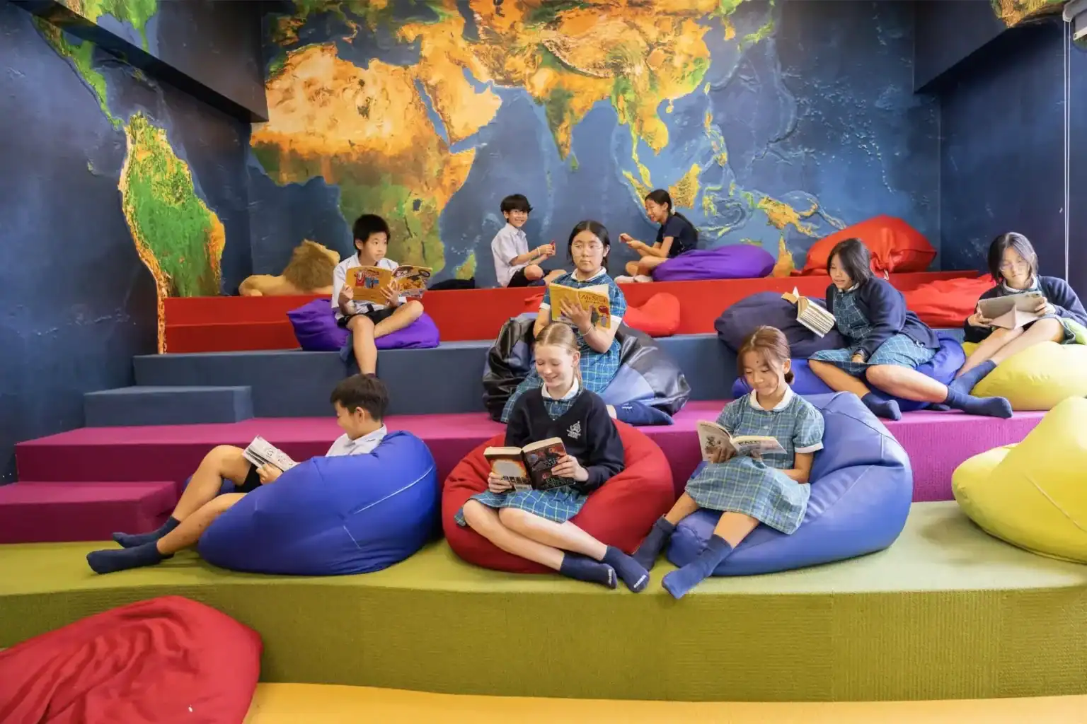 A colorful indoor reading area with children sitting on bean bags and reading books, featuring a wor.