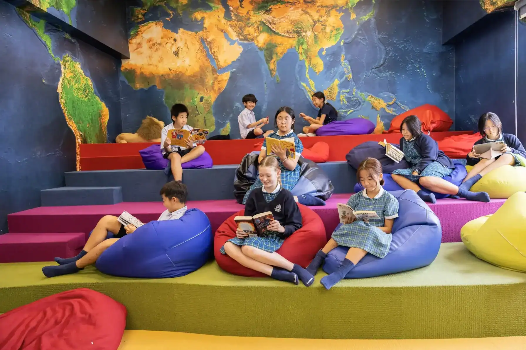 A colorful indoor reading area with children sitting on bean bags and reading books, featuring a wor.