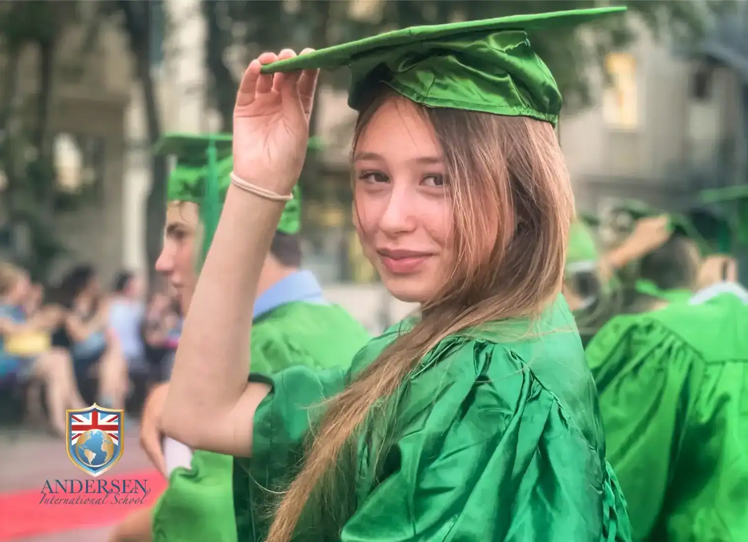 Young female graduate in green cap and gown at outdoor ceremony.