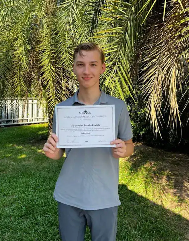 Young student holding a certificate outdoors with palm trees in background.