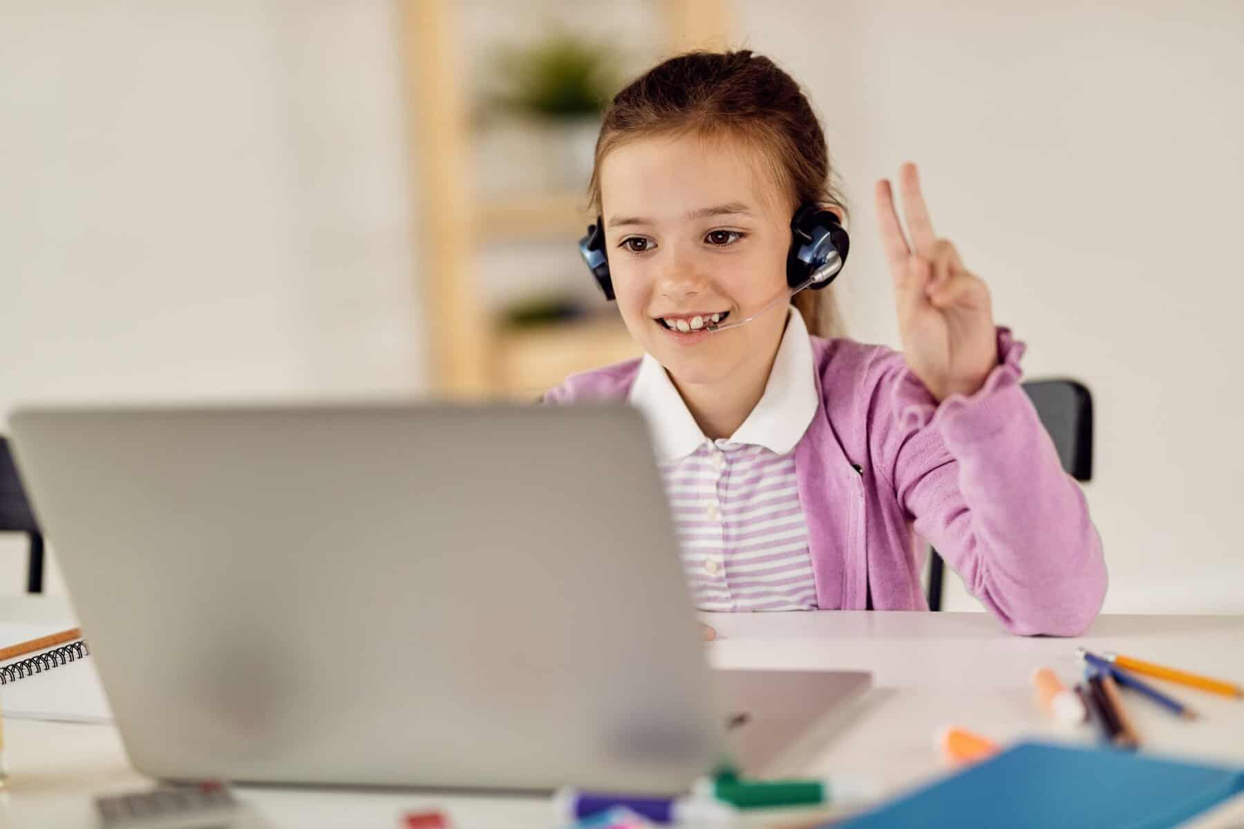 Young girl with headphones smiling and making a peace sign during online learning.