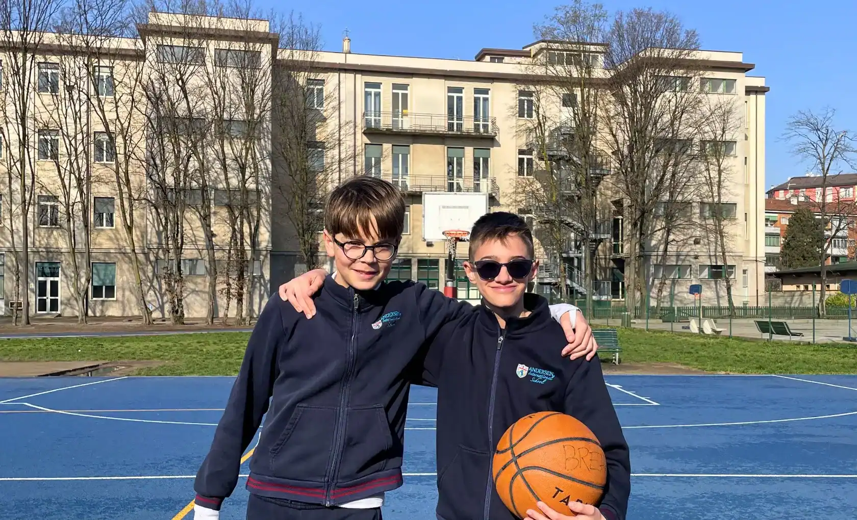 Two boys in school uniforms on a basketball court with school building in background.
