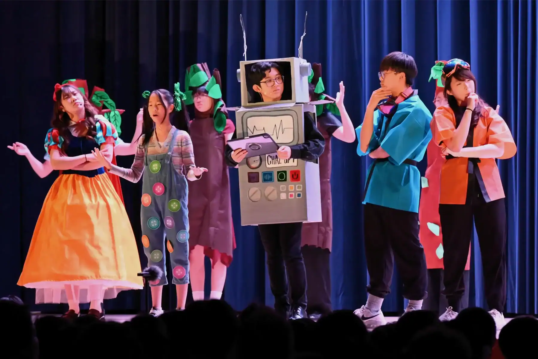 Students perform a school play on stage with colorful costumes and a robot costume.