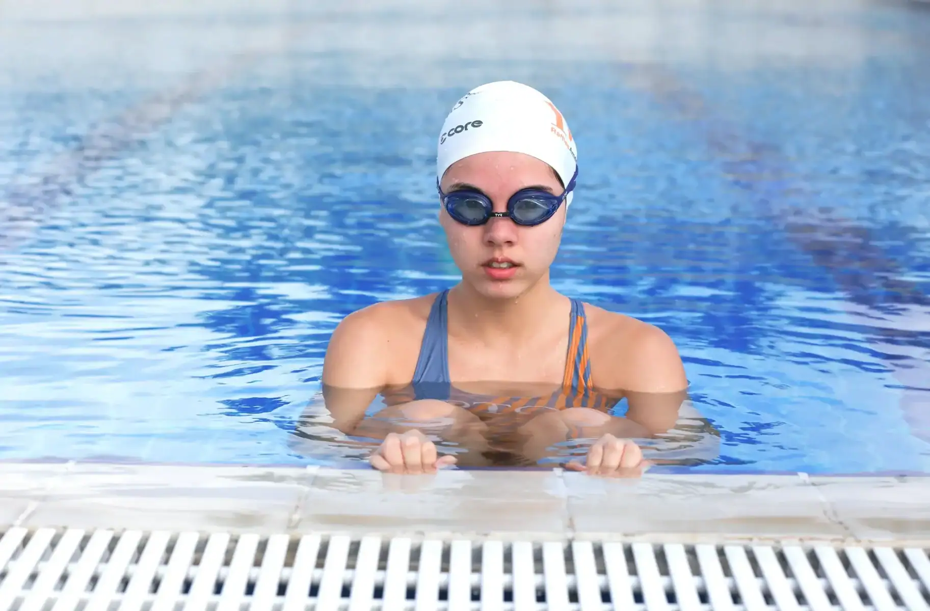 Young girl swimming with goggles and swim cap at the pool edge.