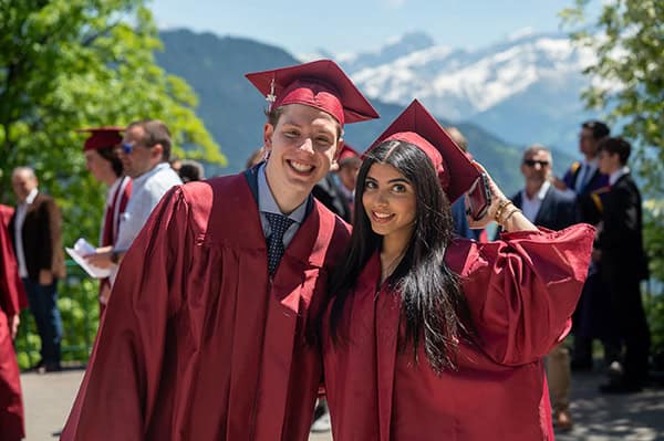 Graduates in maroon caps and gowns celebrating outdoors with mountains in background.