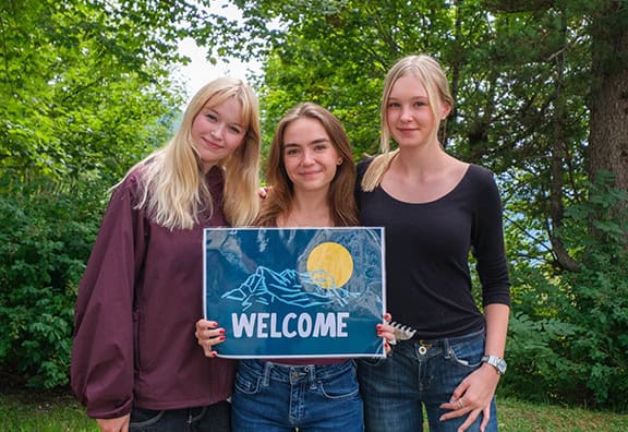 Three young women holding a welcome sign outdoors in a green park.