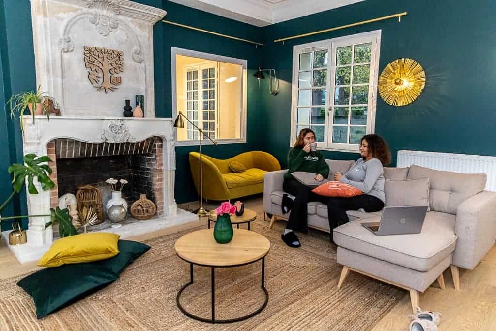 Cozy living room with two women relaxing and chatting on a sofa, bright decor, and large windows.