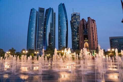City skyline with tall buildings and illuminated fountains at dusk.